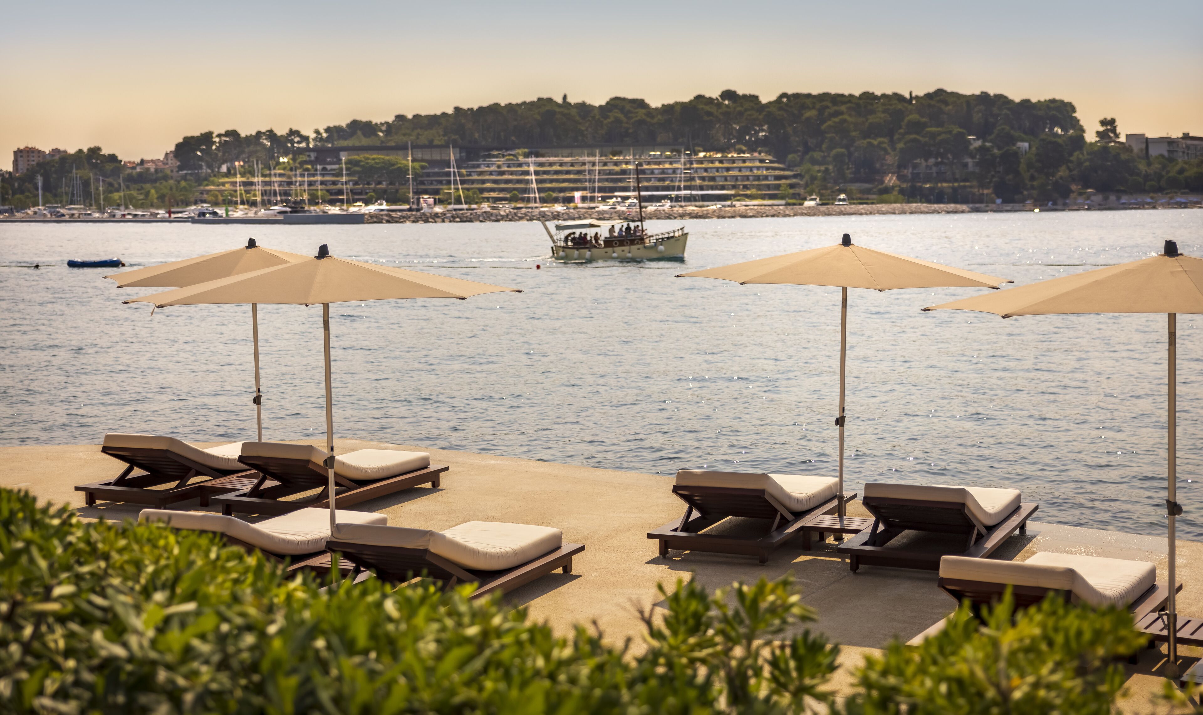 A beachside lounge area with sun loungers and umbrellas, overlooking a calm sea with boats and a distant shoreline.