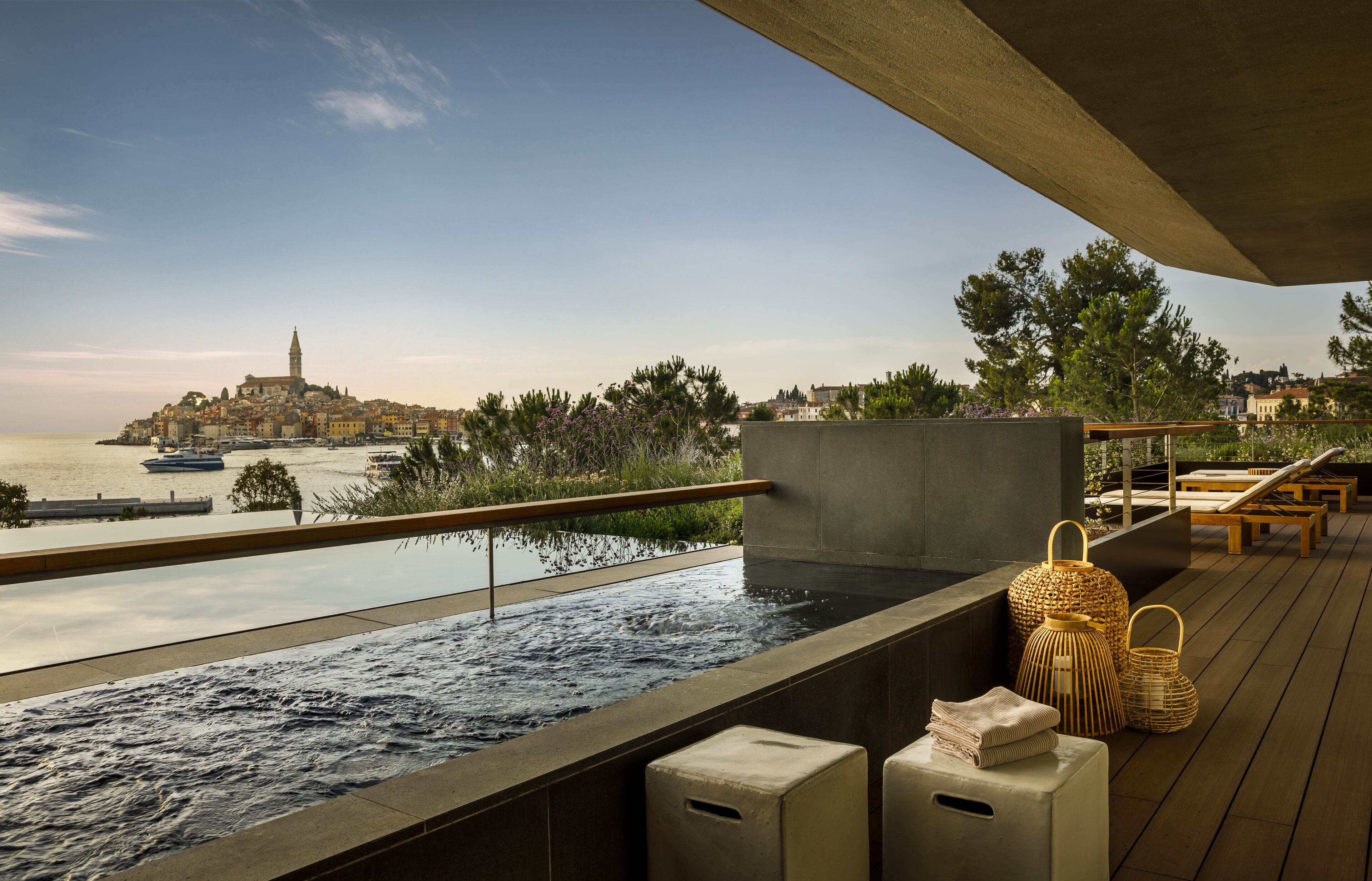 Eine Dachterrasse mit Pool und Blick auf eine Küstenstadt, Holzmöbel und dekorative Laternen.