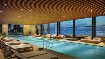 Un espace piscine intérieur moderne, doté de chaises longues et offrant une vue sur le ciel du soir.