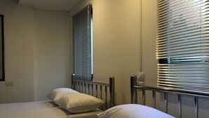 Habitación de hotel con dos camas individuales, cada una con ropa de cama y almohadas blancas, cabeceros metálicos y una ventana con persianas.