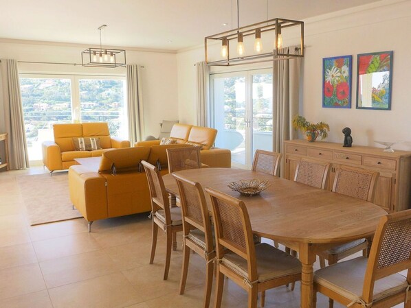 Un salon spacieux avec un canapé jaune, un coin repas comprenant des chaises en bois et une table, et de grandes fenêtres offrant une vue sur l’extérieur.