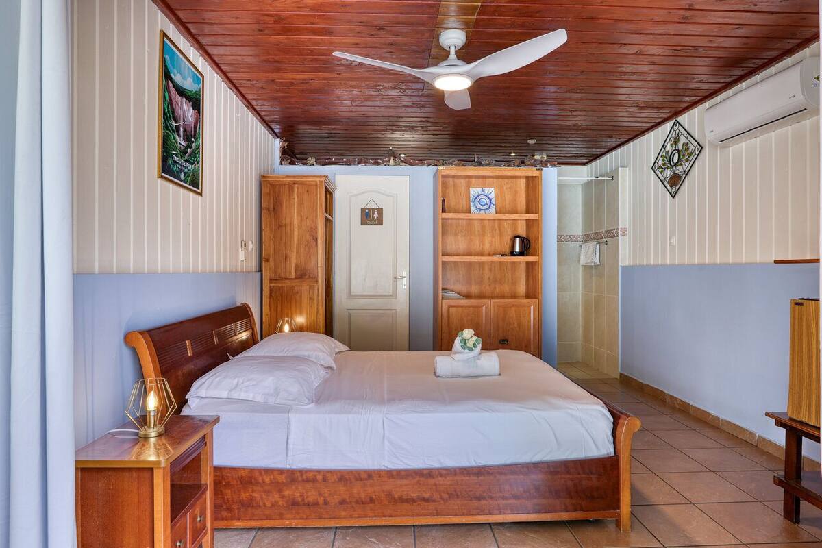 A bedroom with a wooden bed, a ceiling fan, a wooden wardrobe, and a bathroom with a shower.