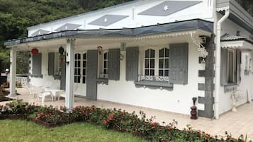 Un edificio blanco con un porche cubierto, un pequeño jardín con flores rojas y montañas al fondo.