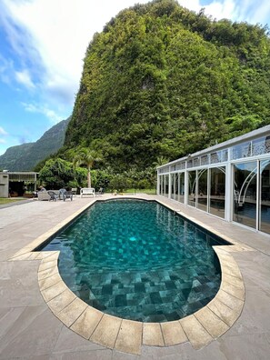Una piscina con borde de piedra, rodeada por un patio con sillas y un cerramiento de vidrio.