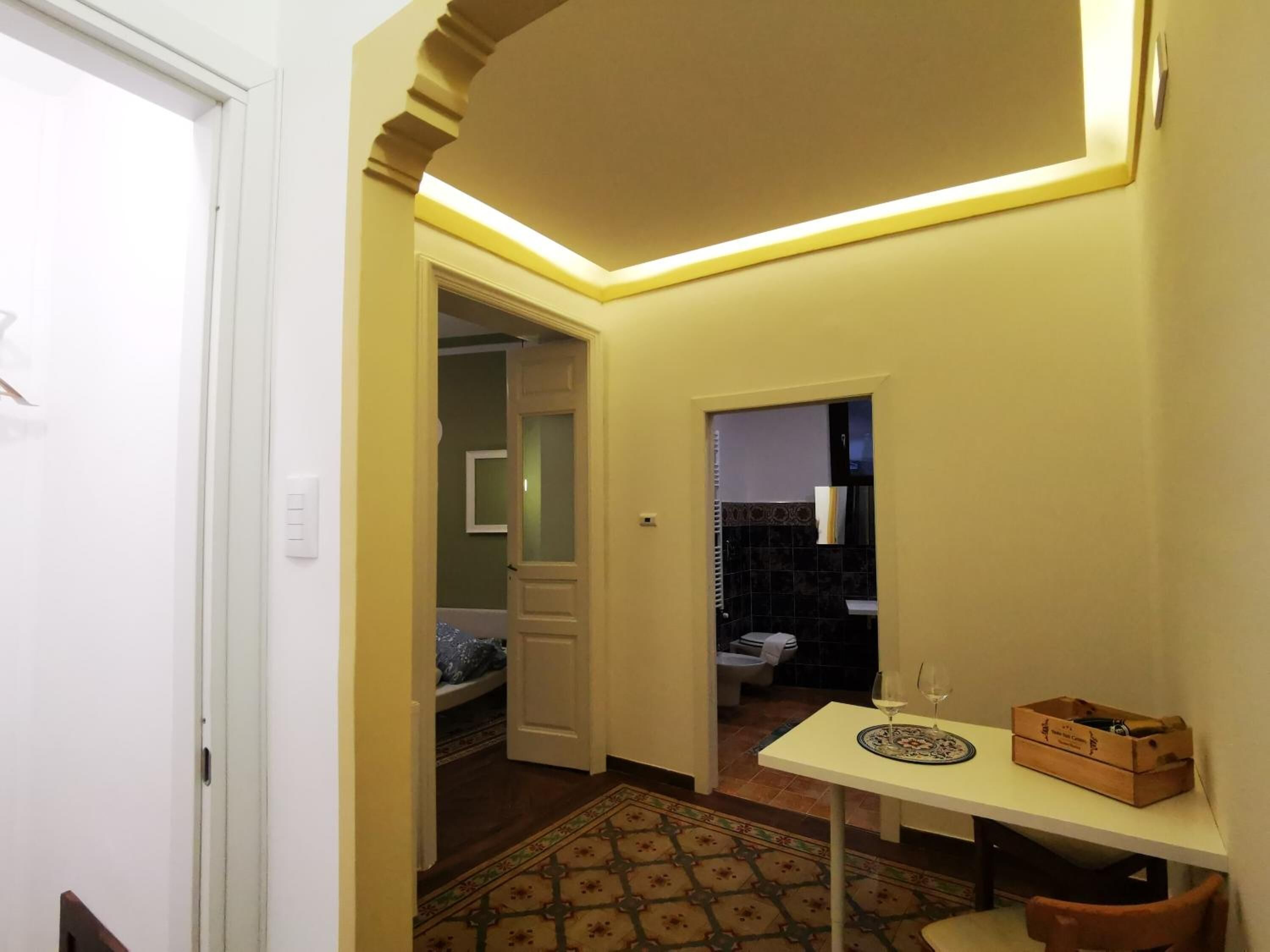 A narrow hallway with a white door on the left, a small table with a glass of wine and a box on the right, and a patterned rug on the floor.
