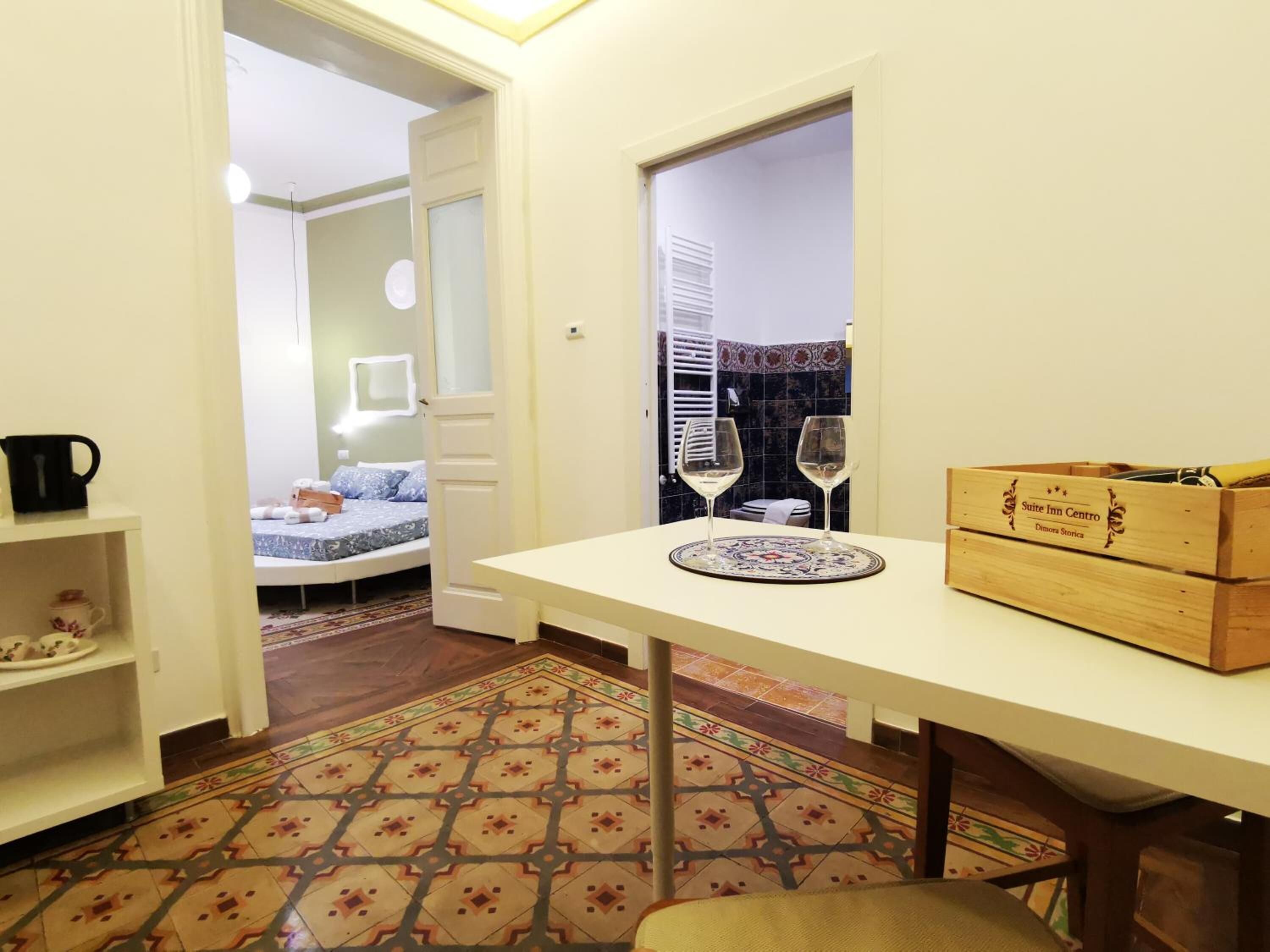 A small, well-lit room with a bed, a table with two wine glasses, and a box on the table.