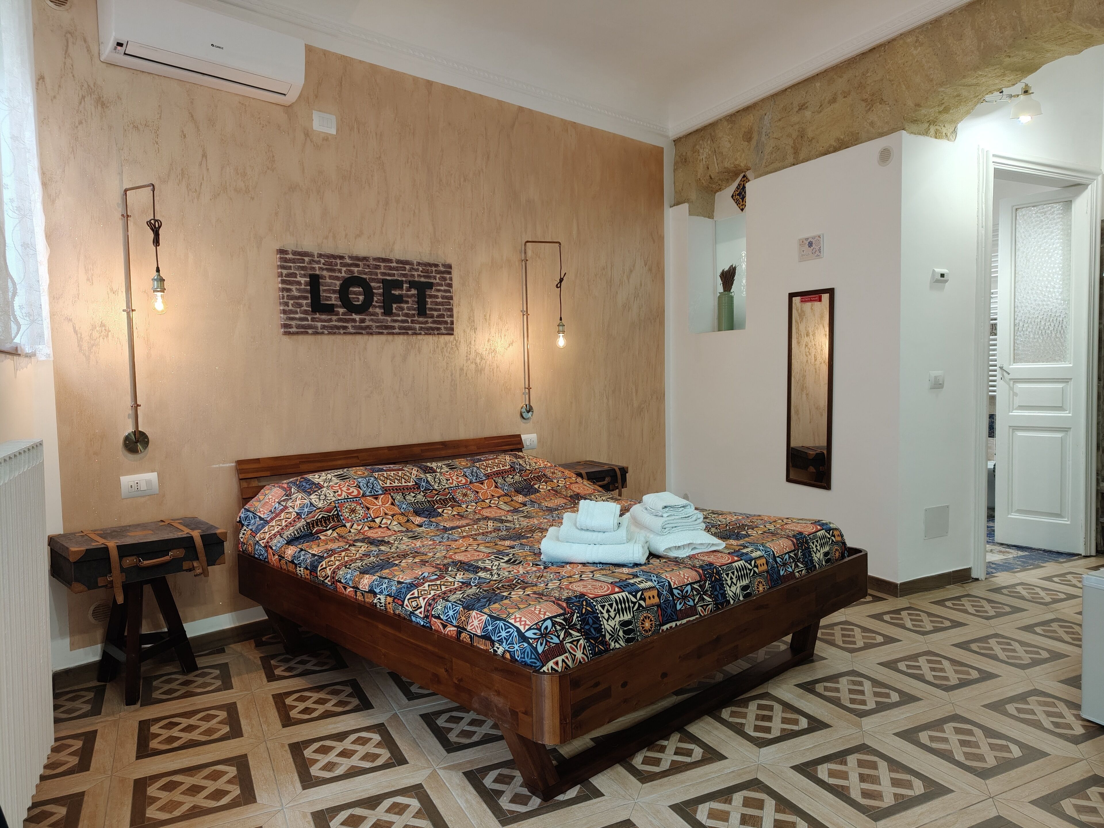 Un dormitorio con edredón estampado, cabecera de madera, aire acondicionado de pared y una mesita de noche pequeña.