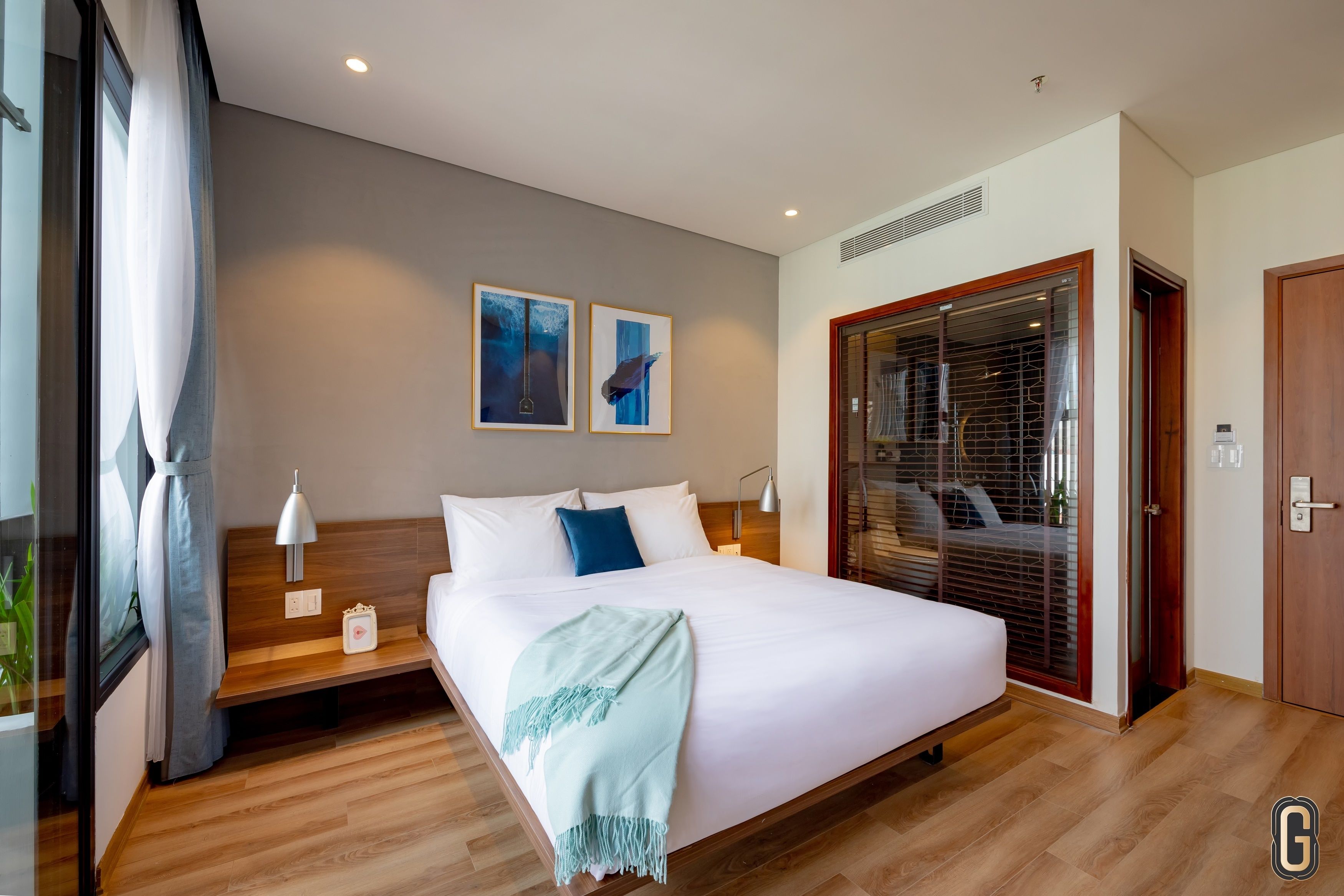 A modern hotel room with a large bed, bedside tables, a nightstand, a window with curtains, and artwork on the wall.