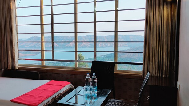 A hotel room with a large window offering a view of a lake and mountains. There is a bed with red pillows, a glass table with water bottles, and a chair. - River Palace (Wai)