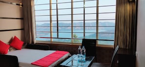 A hotel room with a large window offering a view of a lake and mountains. There is a bed with red pillows, a glass table with water bottles, and a chair. - River Palace (Wai)