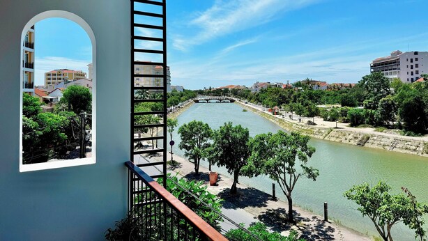 A view from a balcony overlooking a river, with buildings and greenery in the background. - Anriva Hoi An (Da Nang)