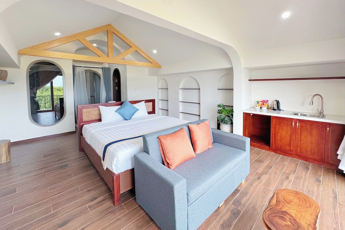 A bedroom with a bed, a sofa, a large window, a kitchen area, and a view of the outdoors.