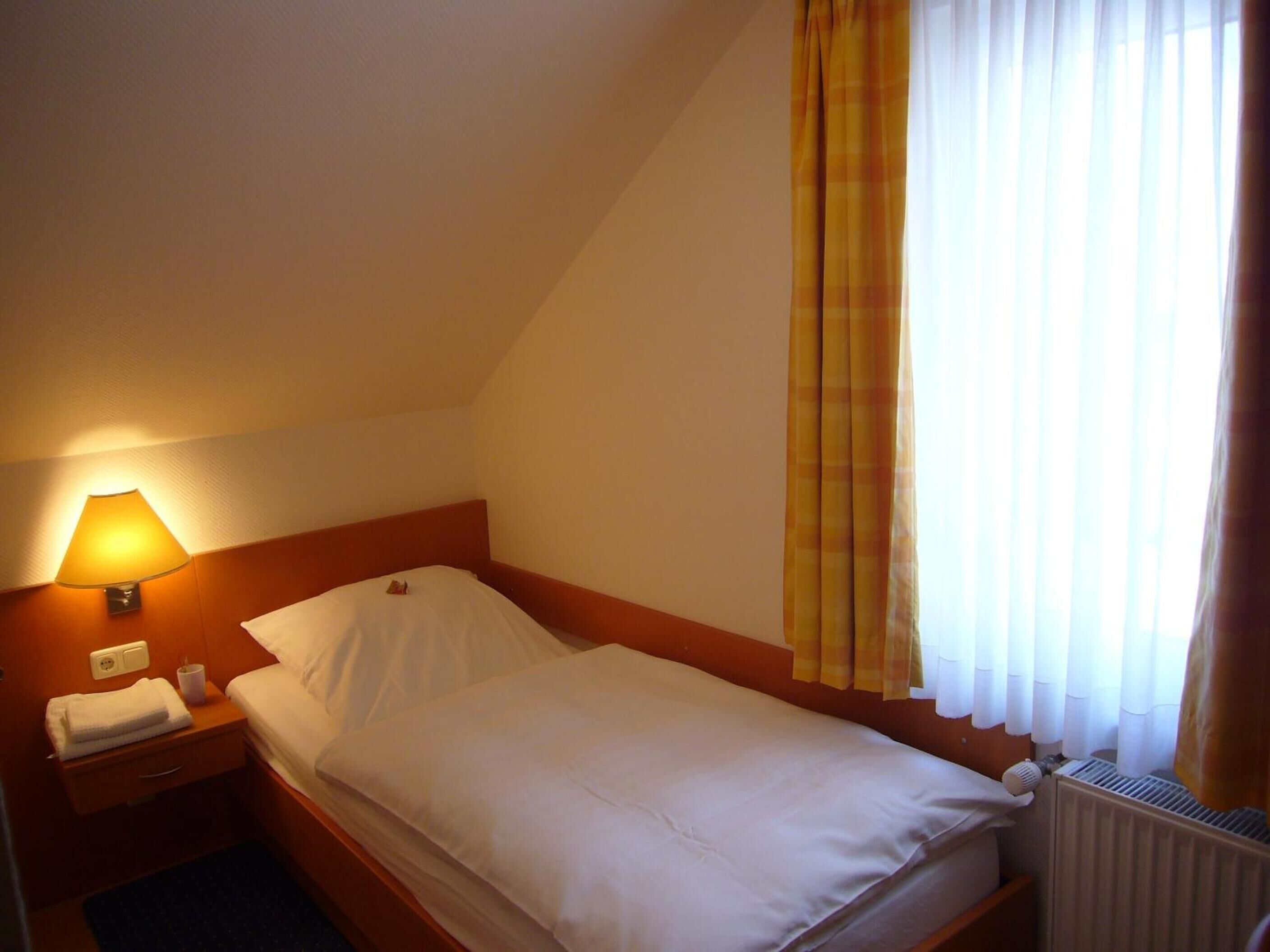 A single bed in a hotel room with a headboard, a bedside lamp, a nightstand, and a window with curtains.