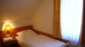 A single bed in a hotel room with a headboard, a bedside lamp, a nightstand, and a window with curtains.
