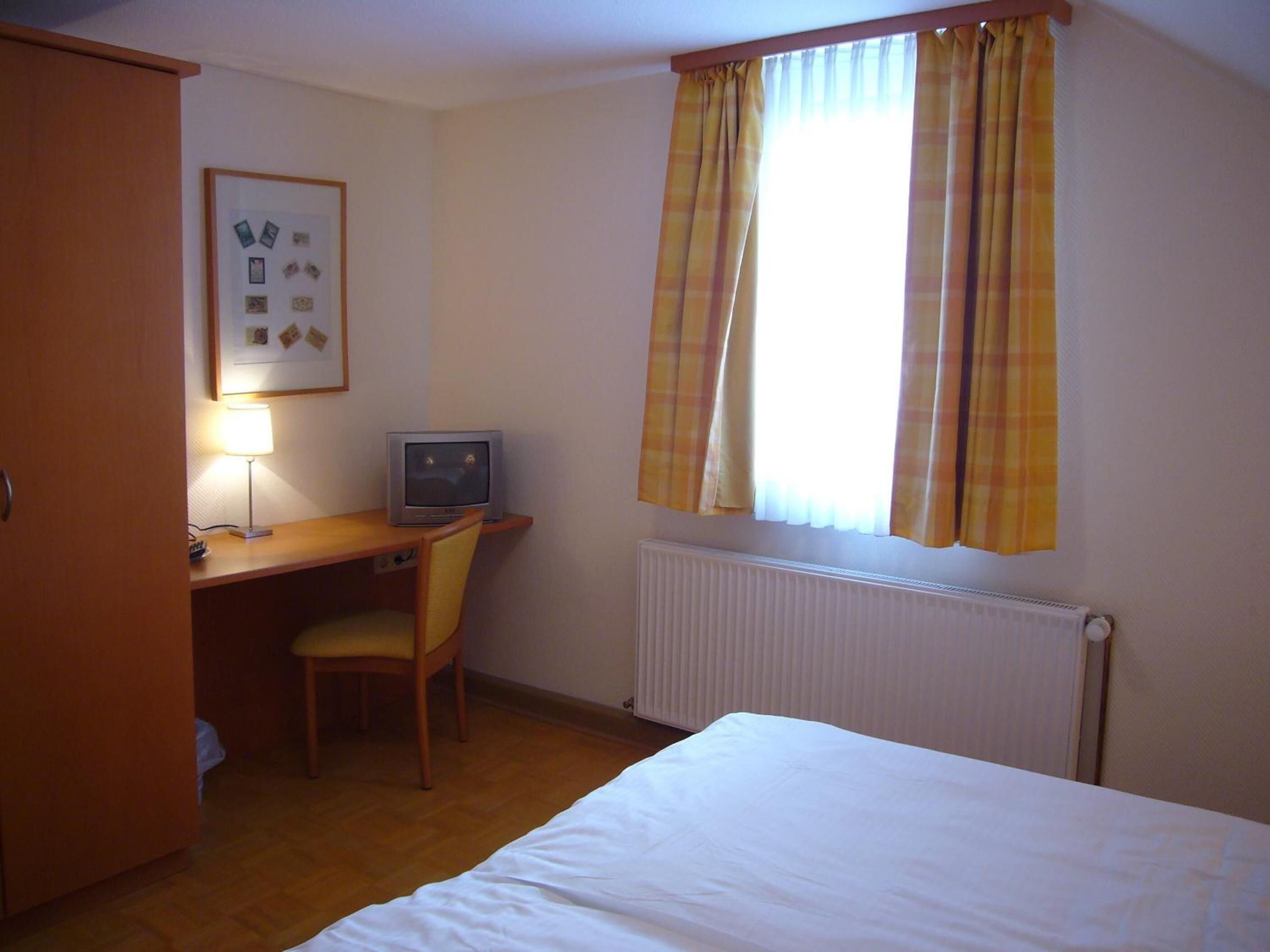 Ein Hotelzimmer mit einem Bett, einem Schreibtisch mit Stuhl, einem Fernseher und einem Fenster mit Vorhängen.