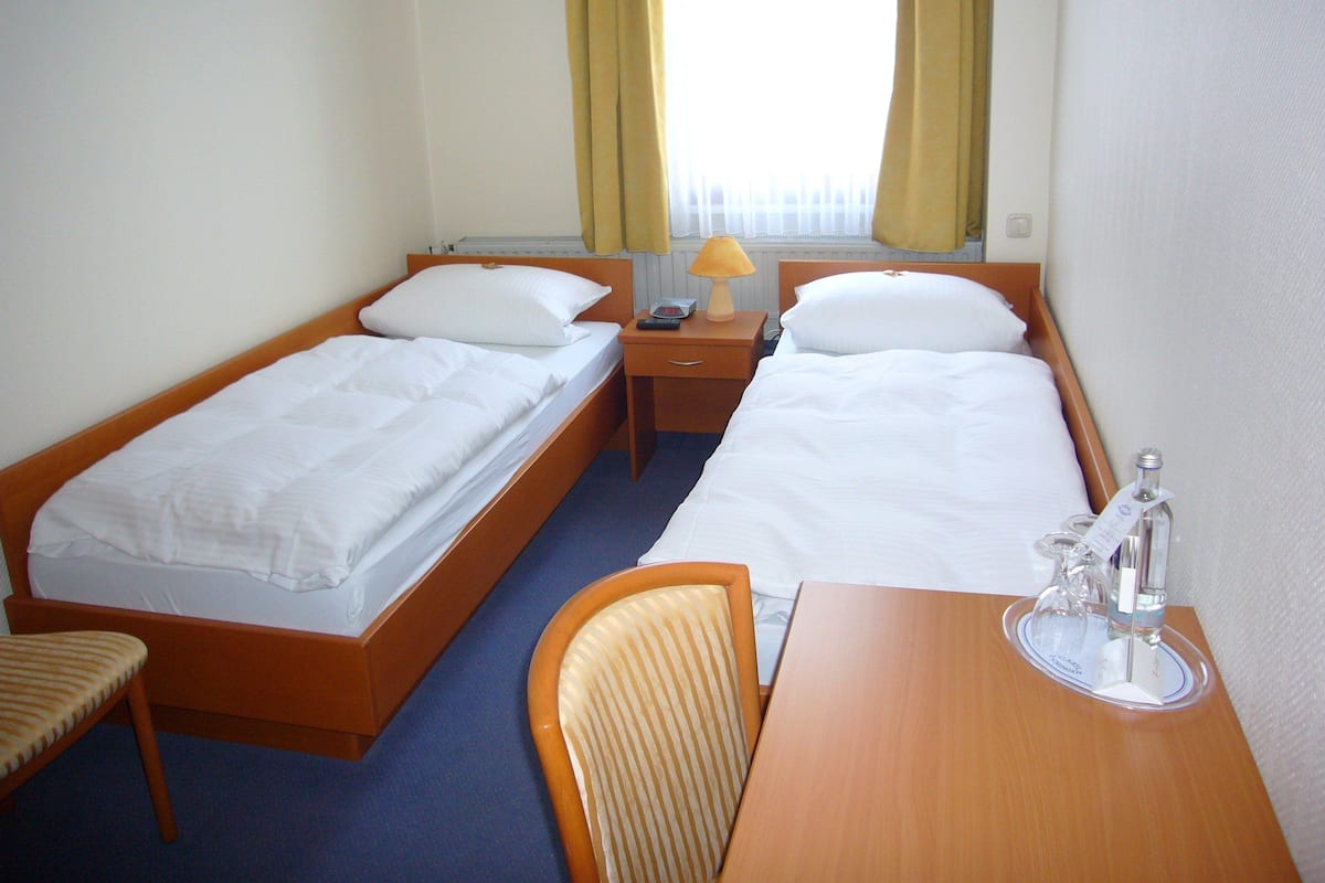 Een hotelkamer met twee eenpersoonsbedden, een houten bureau, een stoel en een klein tafeltje met een fles en een glas.