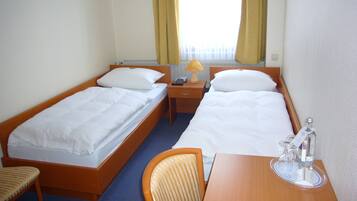 A hotel room with two single beds, a wooden desk, a chair, and a small table with a bottle and glass.