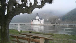 A riverside picnic area with a bench and a small table, overlooking a castle-like building across the river. - Hotel Deutsches Haus (Kaub)