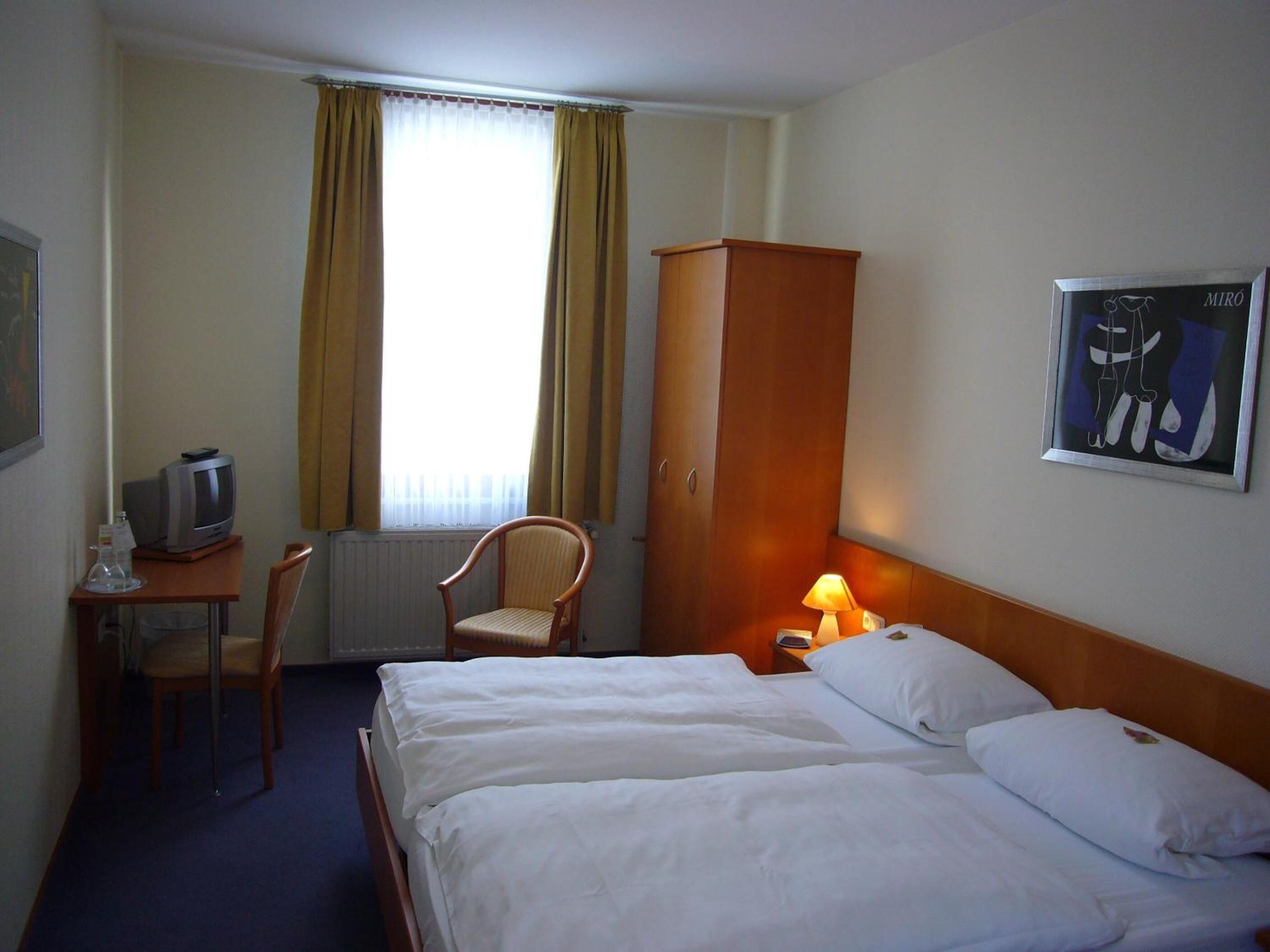 Ett hotellrum med två sängar, en garderob i trä, ett skrivbord med en tv, en stol och ett fönster med gardiner.