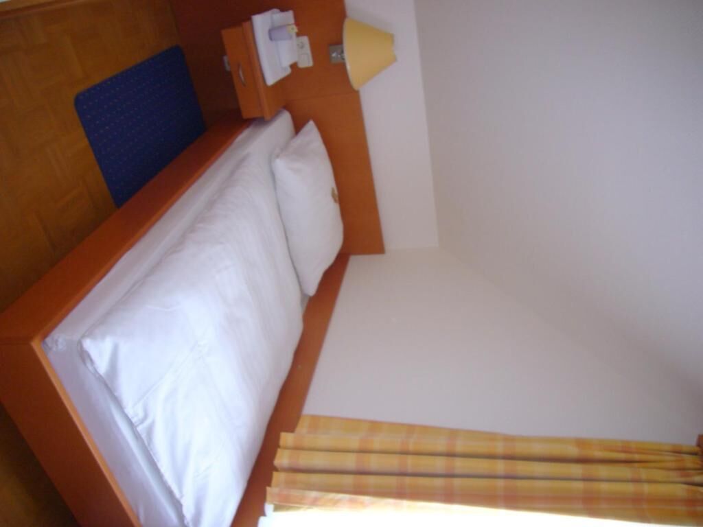 A small, single-bed room with a wooden headboard, a blue headrest, and a window with a plaid curtain.