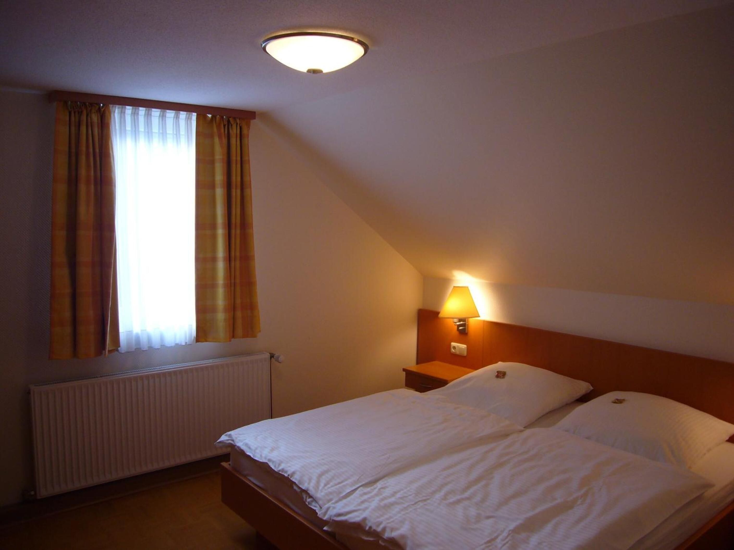 Une chambre d’hôtel avec un lit, une table de chevet, une lampe, une fenêtre avec des rideaux et un radiateur.