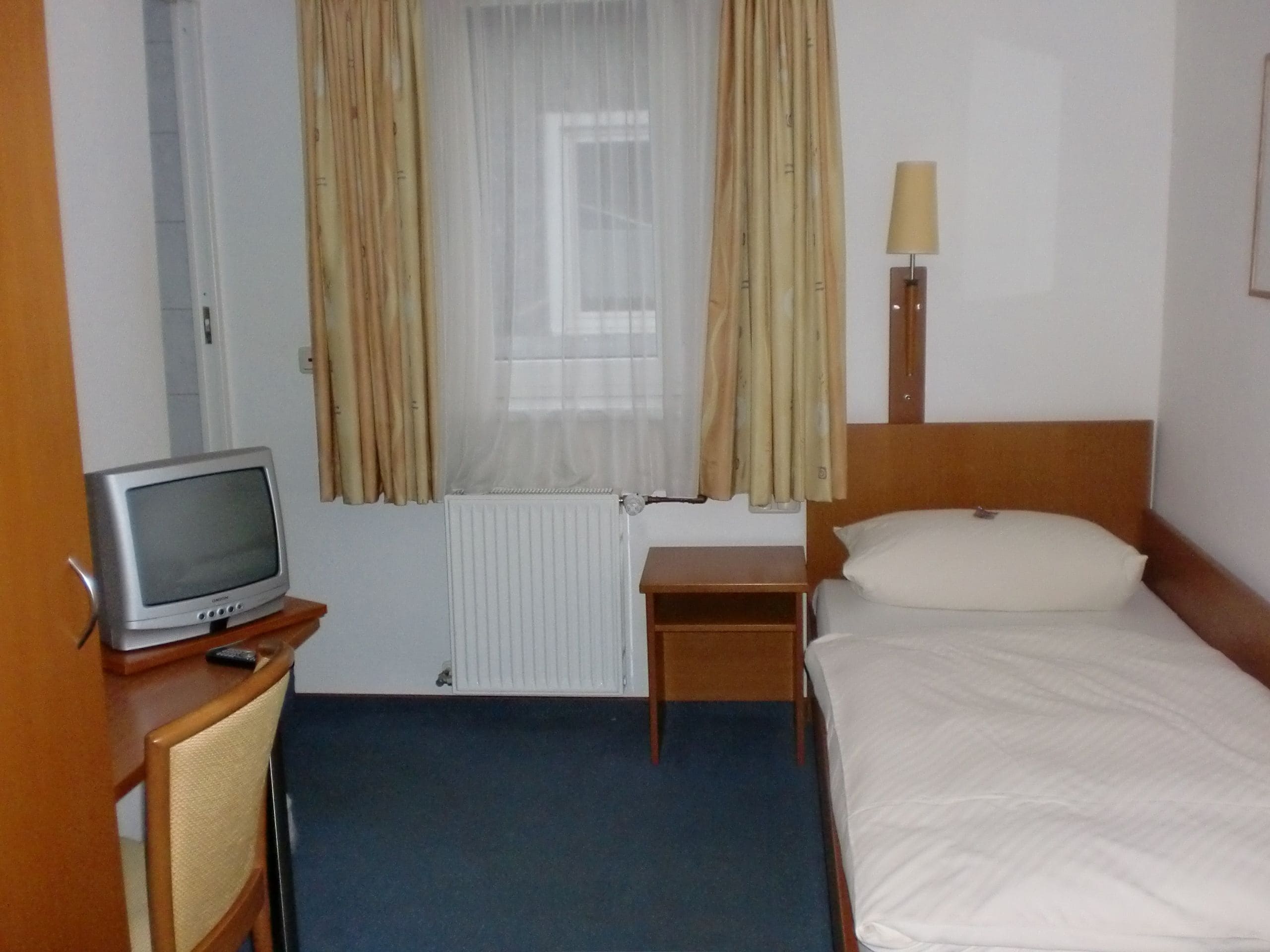 Habitació d'hotel amb un llit individual, una petita taula de nit, una televisió sobre un suport de fusta, una finestra amb cortines i un radiador.