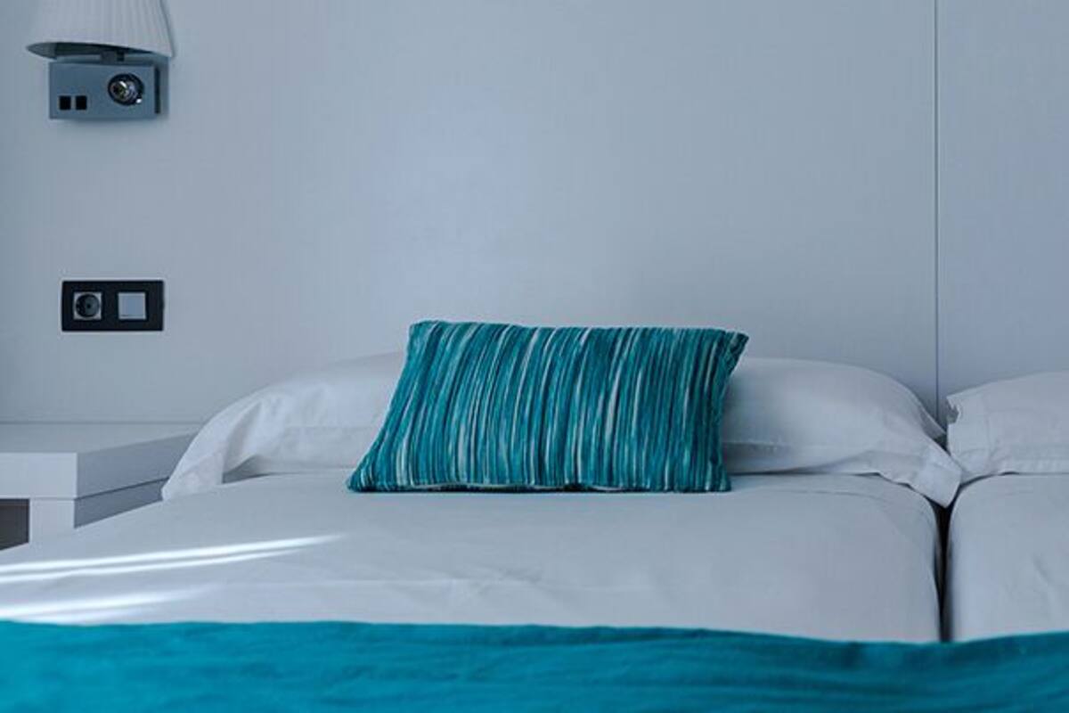 Una cama doble con ropa de cama verde azulada y blanca, dos lámparas de pared y una mesita de noche blanca.