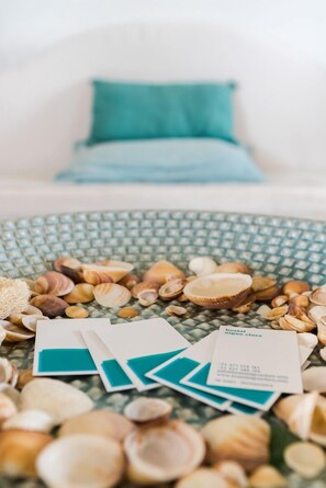 A room with a patterned floor, a teal pillow on a white couch, and scattered seashells and business cards. - Hostal Aigua Clara (Formentera)