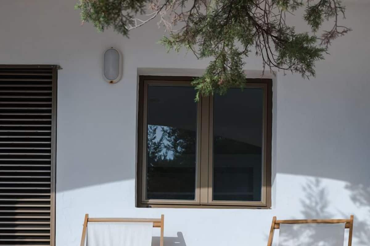 Dos sillones de madera con cojines azules en un patio empedrado, un balde de metal y una ventana con persianas.