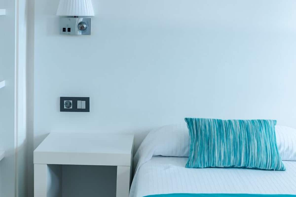 Una habitación de hotel con cama, mesita de noche, lámpara de pared, enchufe y un sombrero colgado en la pared.