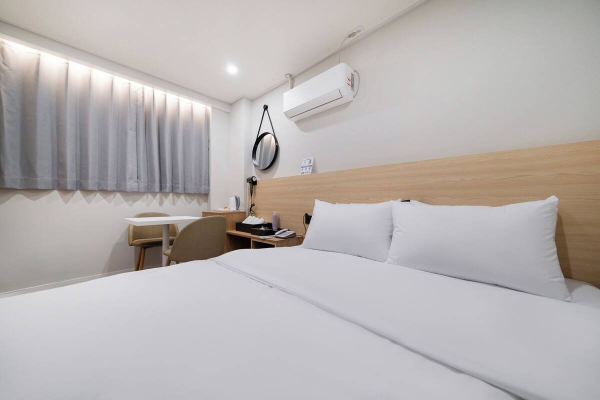 A hotel room with a bed, a desk, a chair, a window with curtains, an air conditioning unit, and a wall-mounted clock.