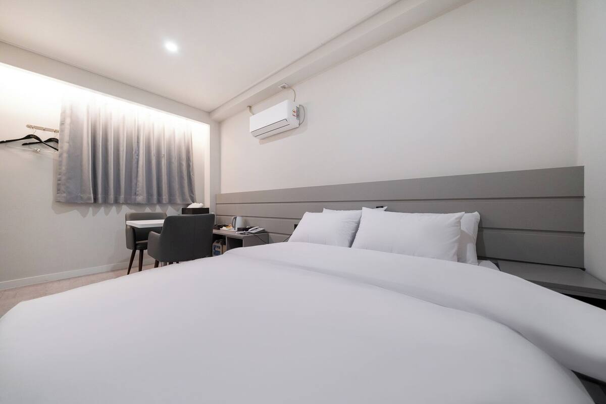 A modern hotel room with a large bed, a desk with a chair, a small table, and a window with curtains.