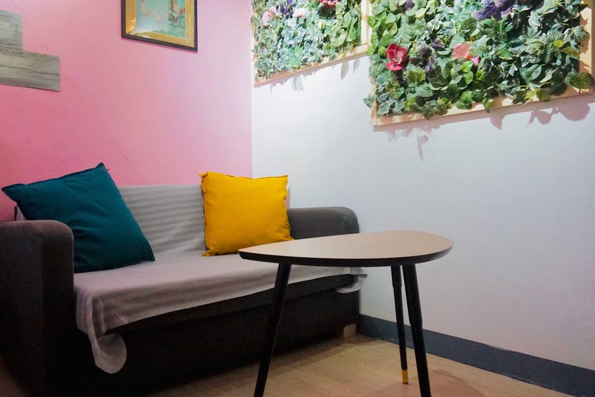 Una habitación con sofá, una mesita, pared rosa, estante de madera y planta colgante.