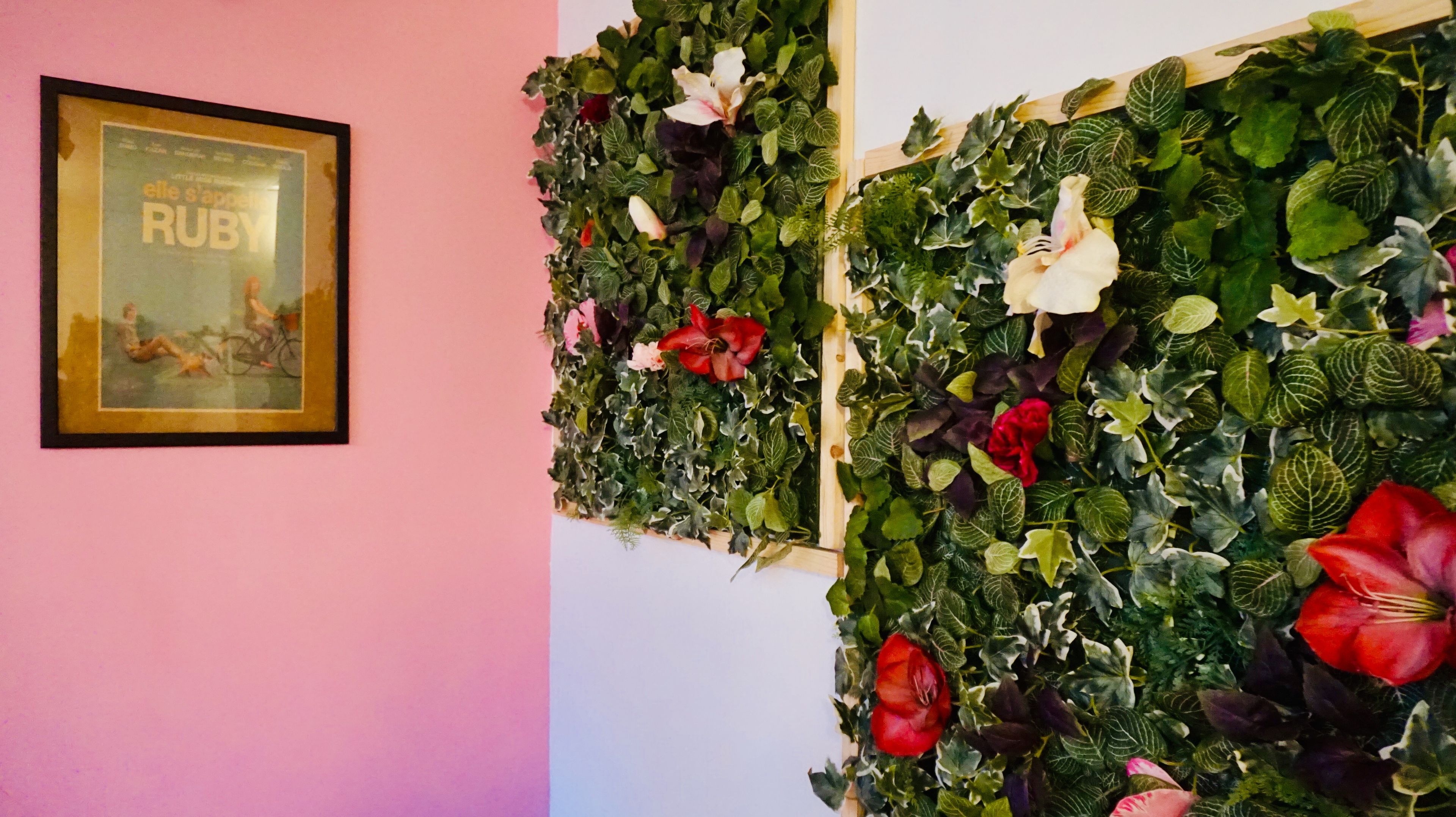 Un cartel de película enmarco sobre una pared rosa y un jardín vertical con diversas plantas y flores.