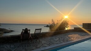 Zona de piscina con suelo de piedra y dos sillas mirando al mar al atardecer.