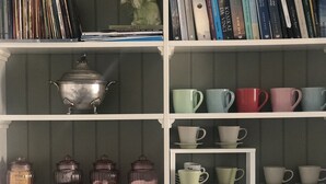 Una cocina con microondas, cafetera y hervidor sobre la encimera, y estantes con libros, tazas y frascos.