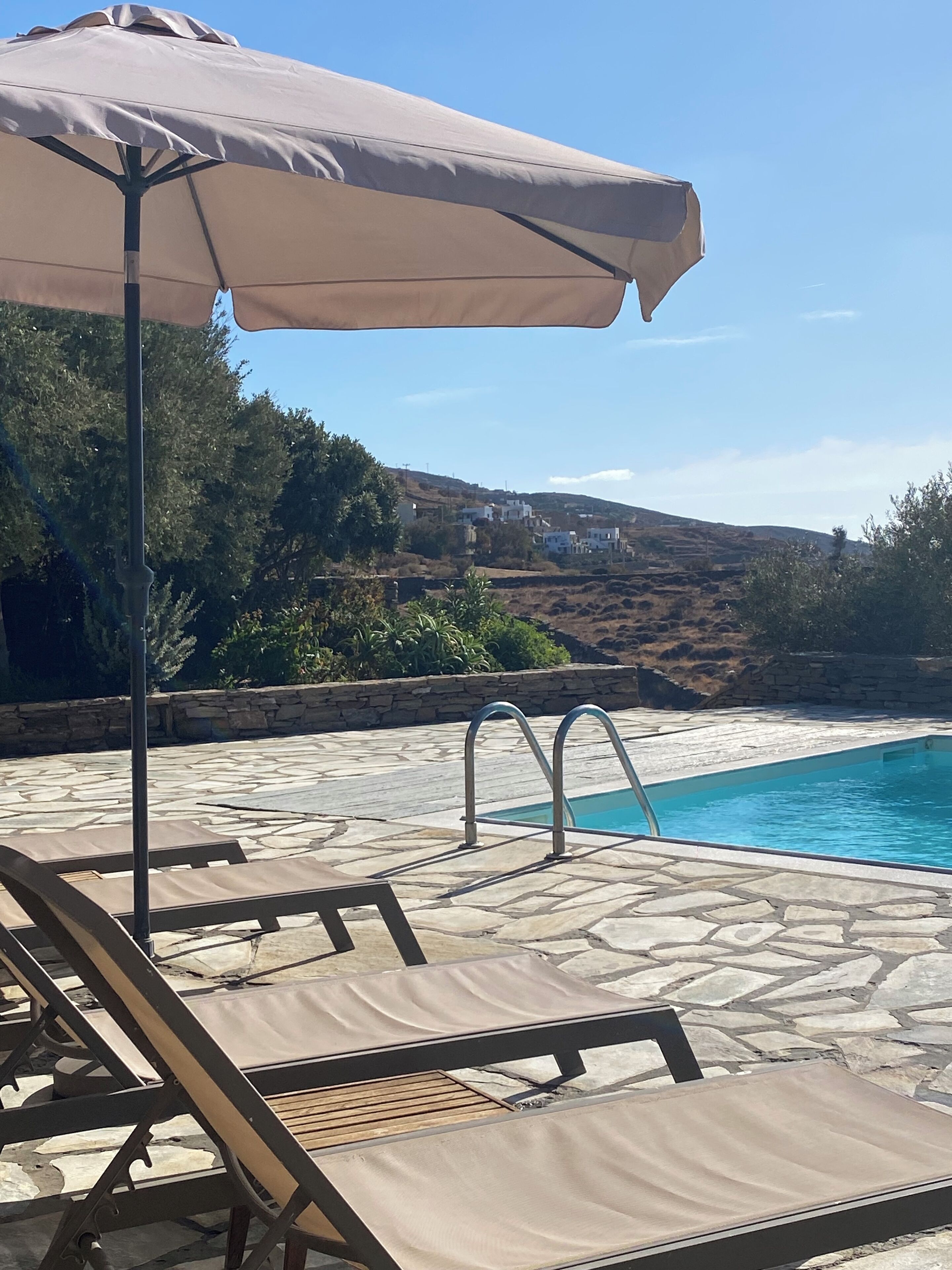 Zona de piscina con sillas de descanso y una sombrilla, suelo de piedra y vistas a una colina con edificios.