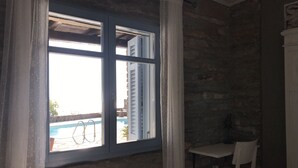 Una habitación con pared de piedra, una ventana con contraventanas blancas, una piscina visible en el exterior y una silla blanca.