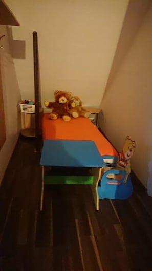 A small, narrow room with a single bed featuring a blue and orange mattress. A teddy bear is placed on the bed, and a small toy figure is on the footboard.