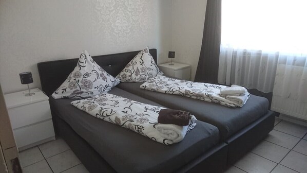 A double bed with floral-patterned bedding and pillows, a bedside table with a lamp, and a window with curtains.