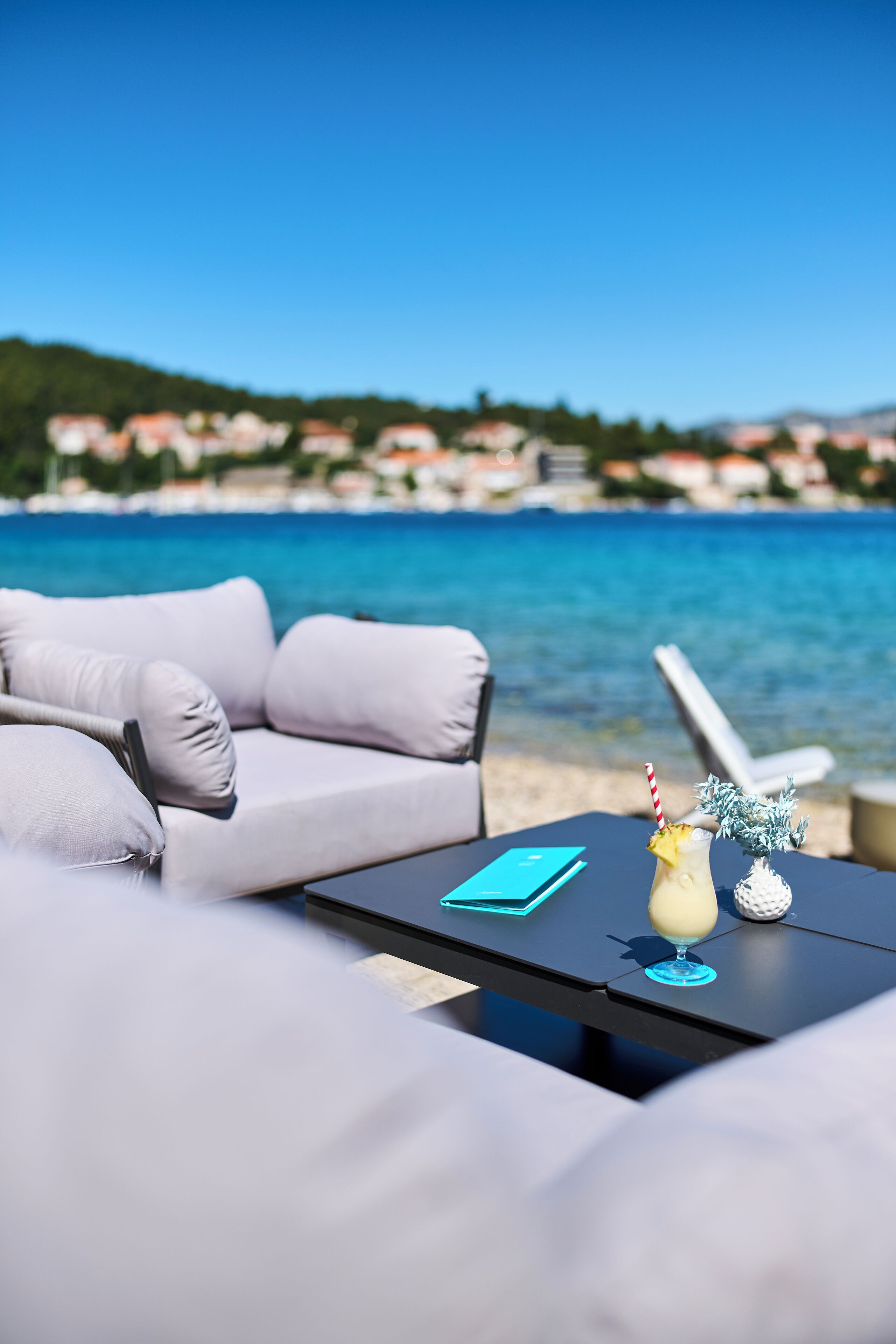 A beachside lounge with a sofa and a table, overlooking a clear blue sea and a distant coastline.