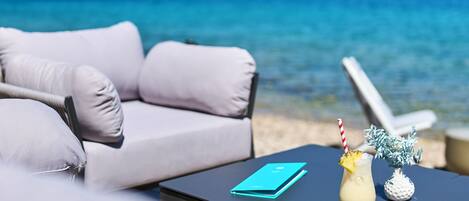A beachside lounge with a sofa and a table, overlooking a clear blue sea and a distant coastline.