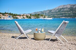 Deux fauteuils de jardin blancs, placés face à une plage de galets, offrent une vue dégagée sur les bateaux et sont encadrés par un paysage montagneux.