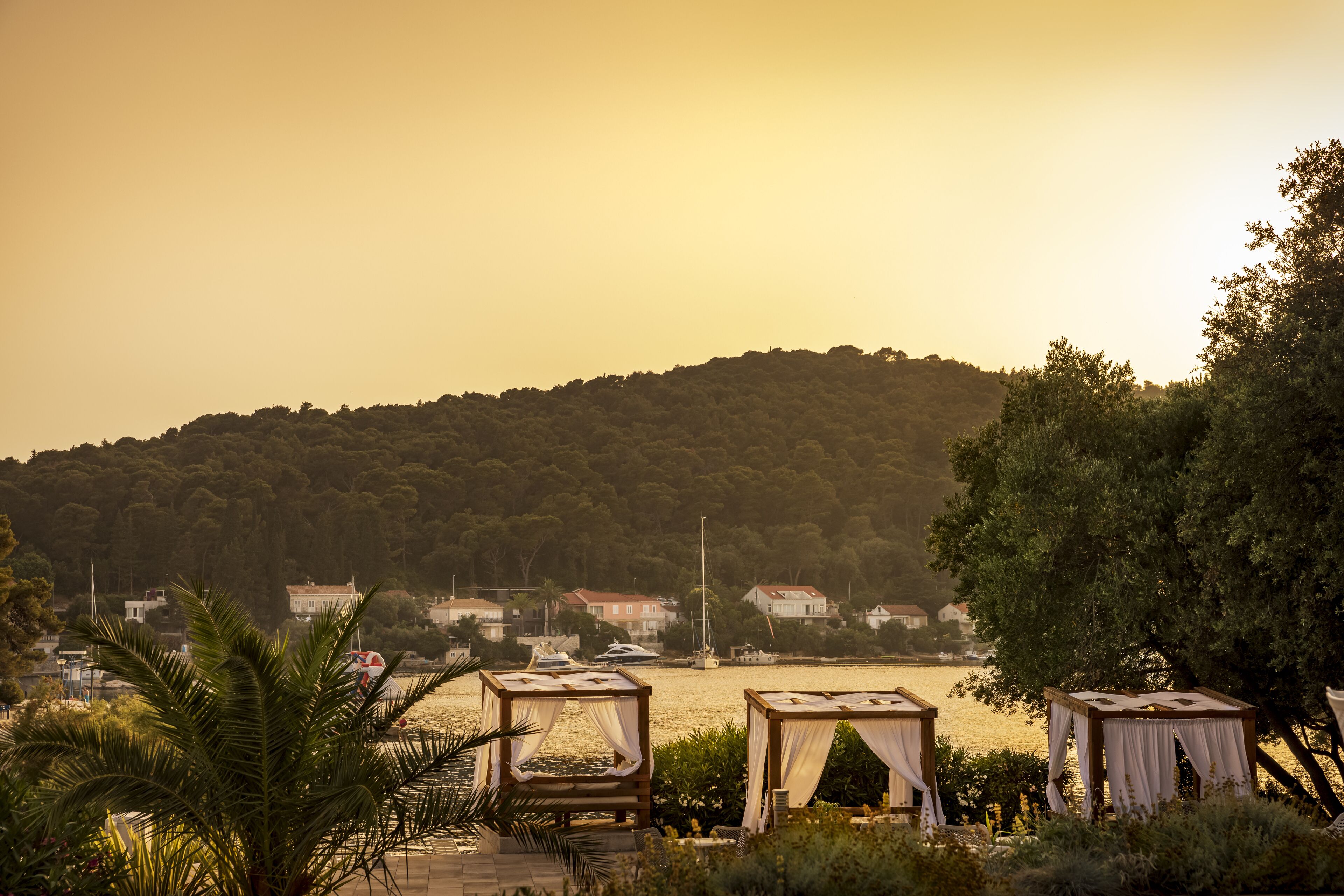 A beachside view with two wooden gazebos, a palm tree, and a distant view of houses and boats on a hill.