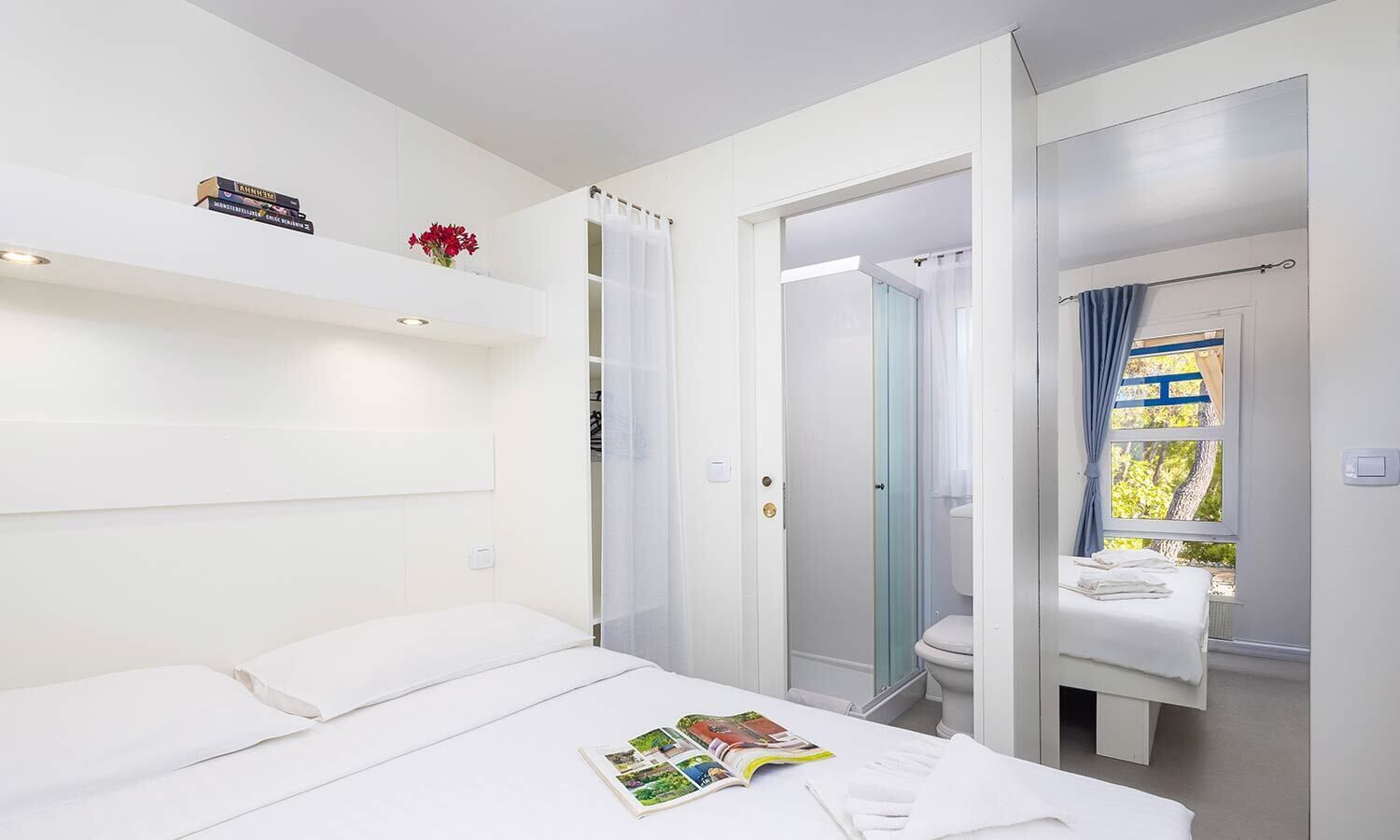 Ett modernt hotellrum med en säng, ett badrum med dusch och ett fönster med utsikt över träd.