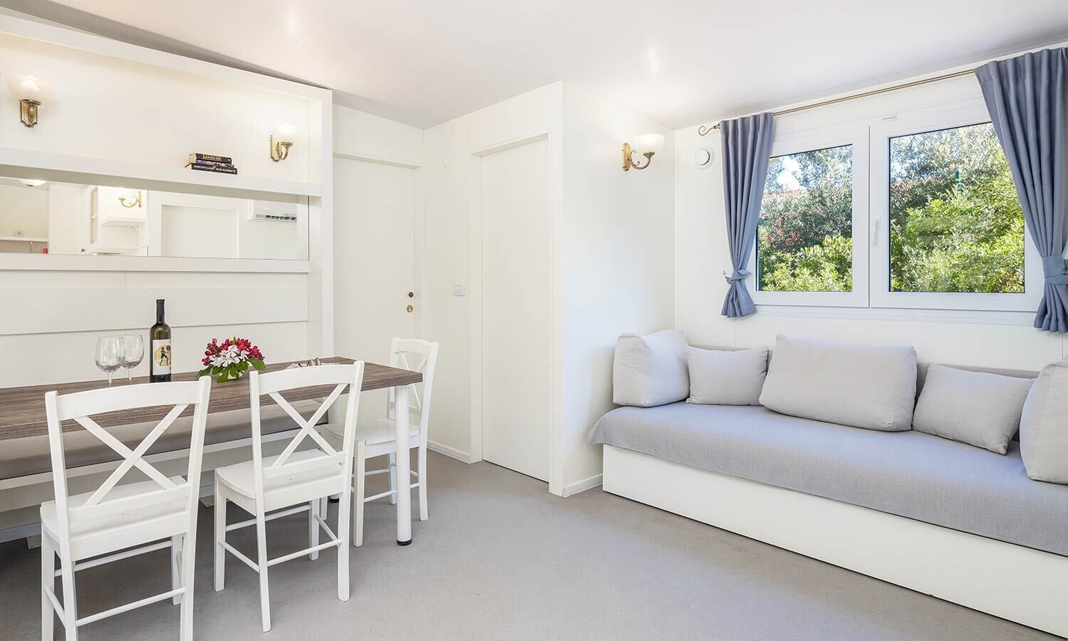 Ein moderner, hell eingerichteter Raum mit Essbereich, der einen weißen Tisch und Stühle, ein graues Sofa mit Kissen und ein Fenster mit Vorhängen umfasst.