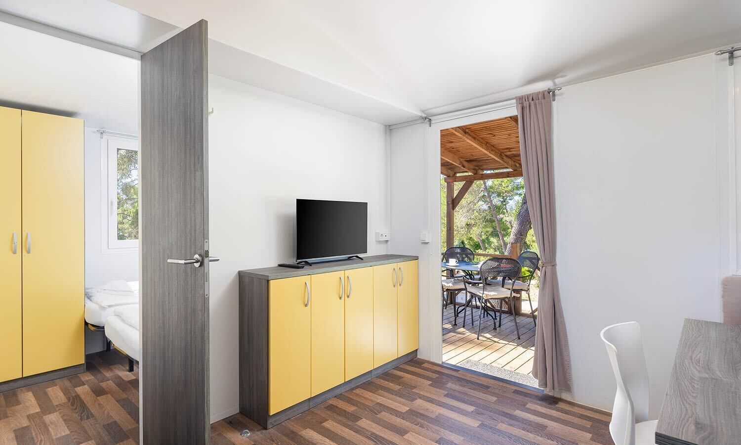 Une chambre moderne avec un lit, un téléviseur posé sur un meuble jaune, et un espace extérieur avec une table et des chaises.