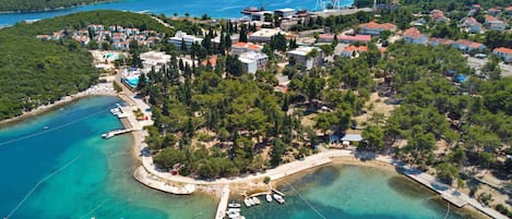 An aerial view of a coastal town with a marina, clear blue waters, and lush green hills in the background.