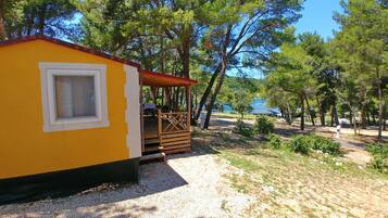 A yellow cabin with a wooden deck and a window, surrounded by trees and a gravel path.