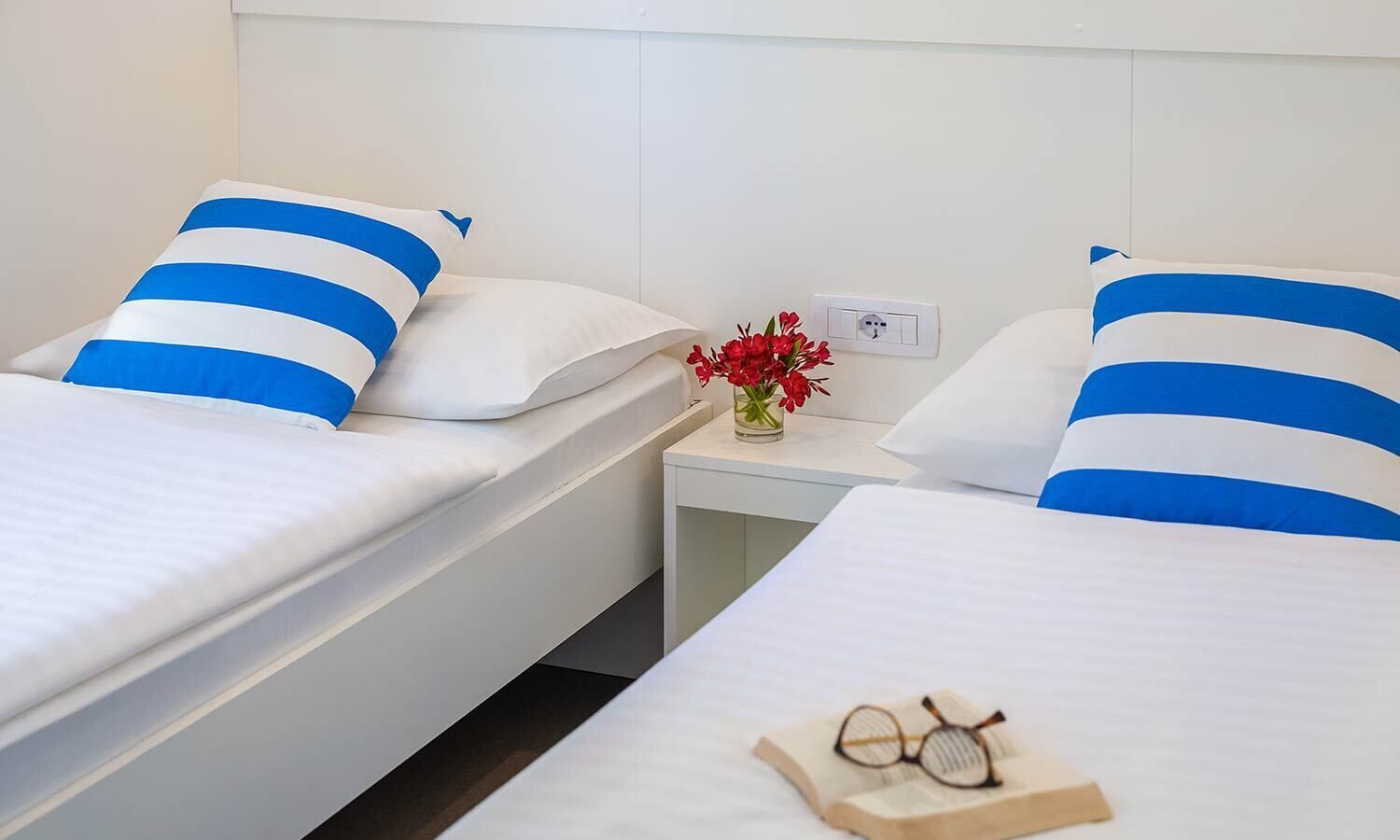 Una habitación de hotel con dos camas individuales, cada una con almohadas a rayas blancas y azules, una mesita de noche con un jarrón de flores rojas y un par de gafas sobre un libro.